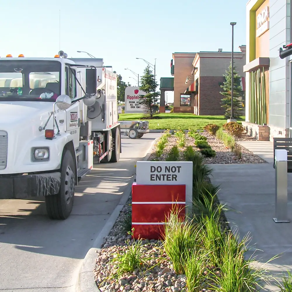 A white truck is parked in a drive-thru lane next to a DO NOT ENTER sign, with buildings, grass, and landscaping visible in the background on a sunny day.