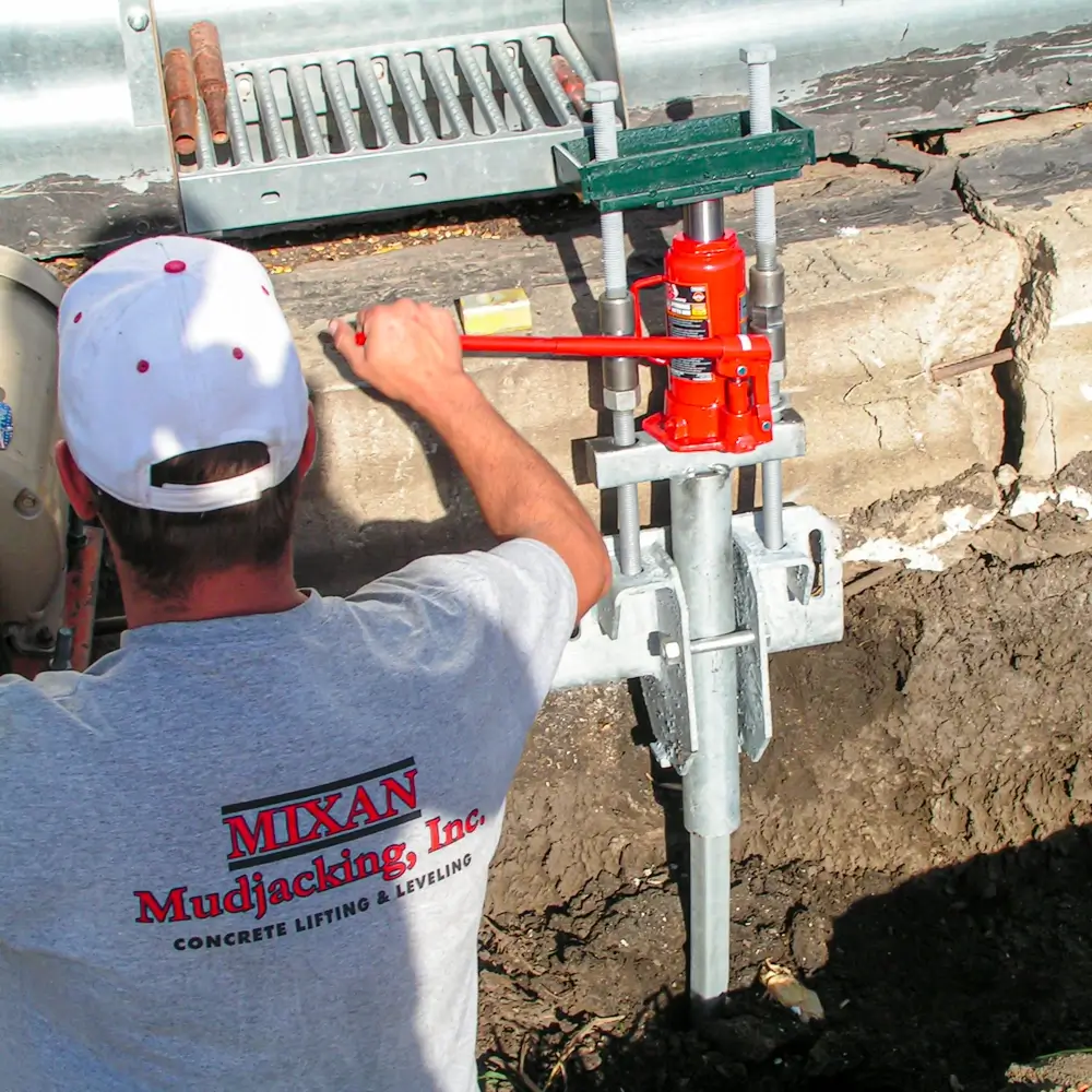 A worker uses a hydraulic jack and metal support to repair and level a cracked concrete foundation near a roadside. He wears a white shirt with Mixan Mudjacking Inc. printed on the back and a white cap. - Mixan Mudjacking A worker uses a hydraulic jack and metal support to repair and level a cracked concrete foundation near a roadside. He wears a white shirt with Mixan Mudjacking Inc. printed on the back and a white cap.