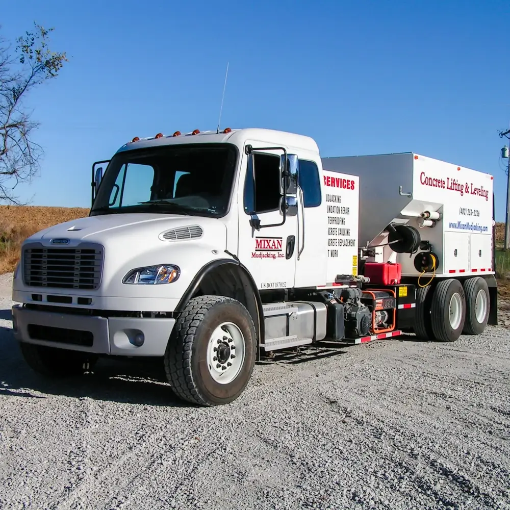 A white truck with Mixan Mudjacking, Inc. signage is parked on gravel. The truck has equipment for concrete lifting and leveling, with details and services listed on the large back section.