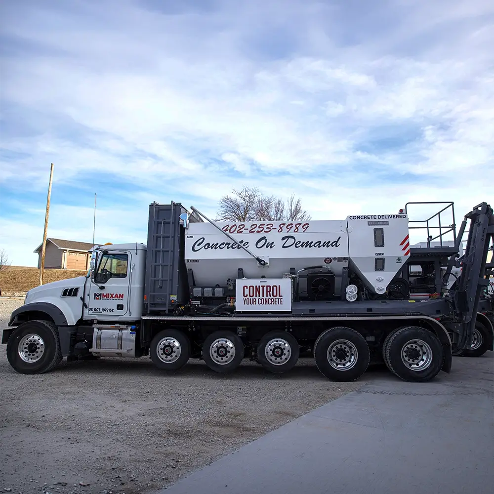 A large concrete mixer truck labeled Concrete On Demand and Control Your Concrete is parked on a gravel surface near a house under a partly cloudy sky. - Mixan Mudjacking A large concrete mixer truck labeled Concrete On Demand and Control Your Concrete is parked on a gravel surface near a house under a partly cloudy sky.