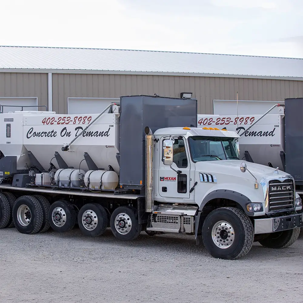 A white Mack concrete mixer truck with Concrete On Demand and a phone number on the side is parked on gravel in front of a metal building.