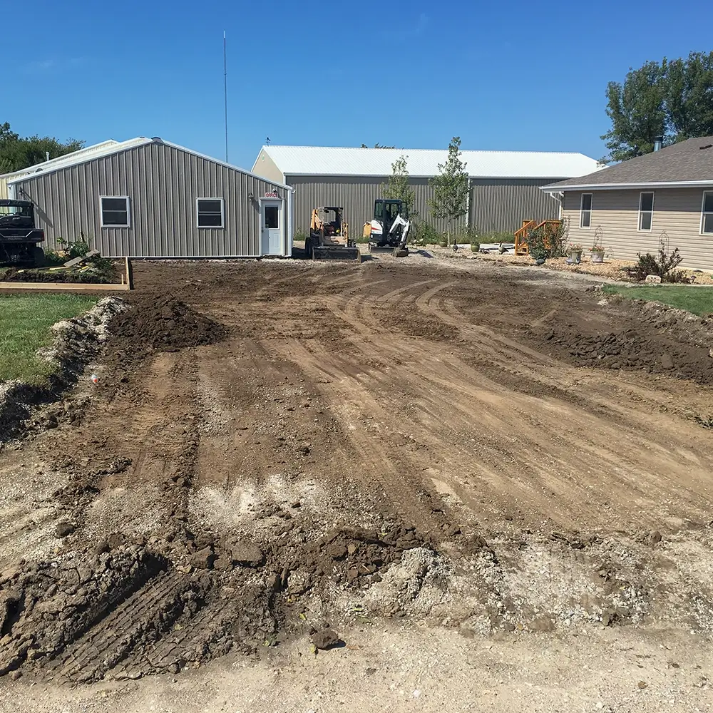 A cleared dirt lot with tire tracks, bordered by a grassy edge, sits between a metal building and a tan house under a blue sky. Construction equipment is visible in the background. - Mixan Mudjacking A cleared dirt lot with tire tracks, bordered by a grassy edge, sits between a metal building and a tan house under a blue sky. Construction equipment is visible in the background.
