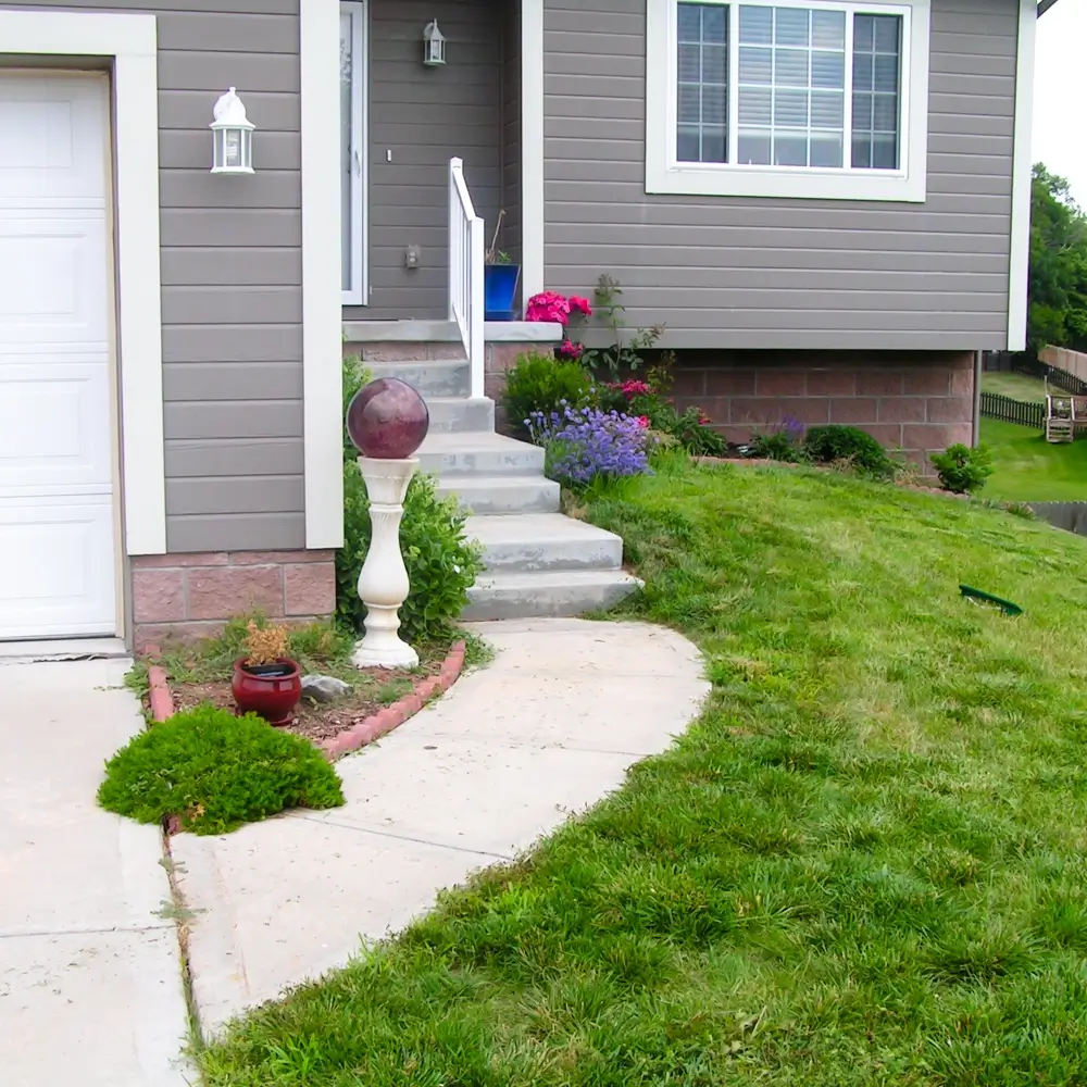 Front yard of a modern house with gray siding, white trim, a concrete walkway, trimmed grass, potted plants, a garden bed with flowers, and steps leading to the front porch.