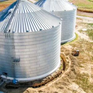 A large metal grain bin sits on a farm surrounded by freshly dug earth, with a small skid-steer loader parked nearby and another bin partially visible in the background.