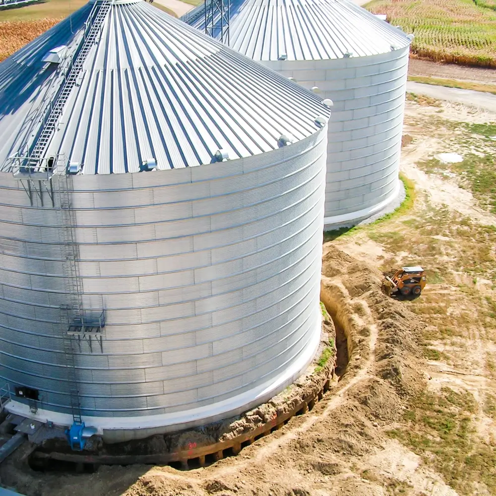 A large metal grain bin sits on a farm surrounded by freshly dug earth, with a small skid-steer loader parked nearby and another bin partially visible in the background.