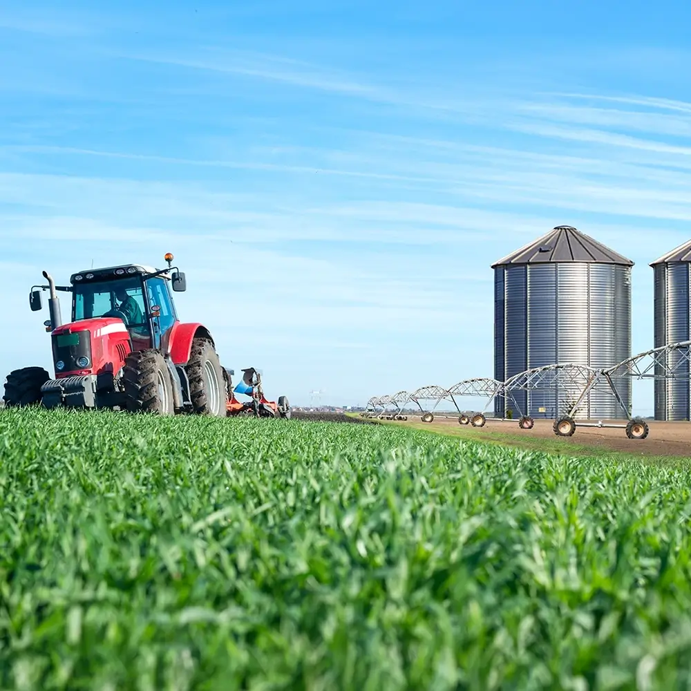 A red tractor works in a green field near large metal silos under a blue sky. An irrigation system extends across the field, indicating active farming operations.