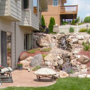 A backyard features a small patio with a chair and a covered fire pit. Behind it, a rocky landscaped slope with a cascading waterfall leads up to a fenced yard and neighboring house. Grass borders the patio area.