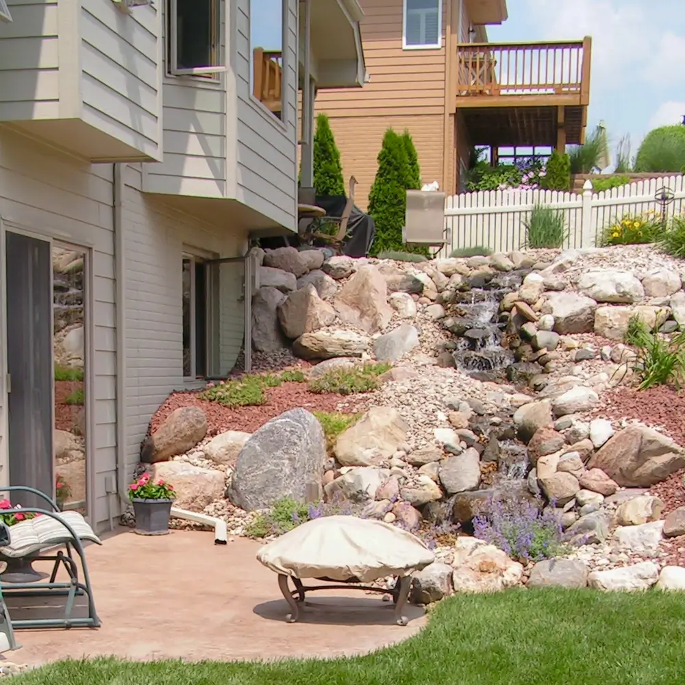 A backyard features a small patio with a chair and a covered fire pit. Behind it, a rocky landscaped slope with a cascading waterfall leads up to a fenced yard and neighboring house. Grass borders the patio area. - Mixan Mudjacking A backyard features a small patio with a chair and a covered fire pit. Behind it, a rocky landscaped slope with a cascading waterfall leads up to a fenced yard and neighboring house. Grass borders the patio area.
