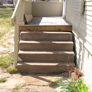 Concrete steps lead up to a porch, but there is no door at the top; the steps end at a blank wall of the house. Some grass and plants grow around the base of the steps.