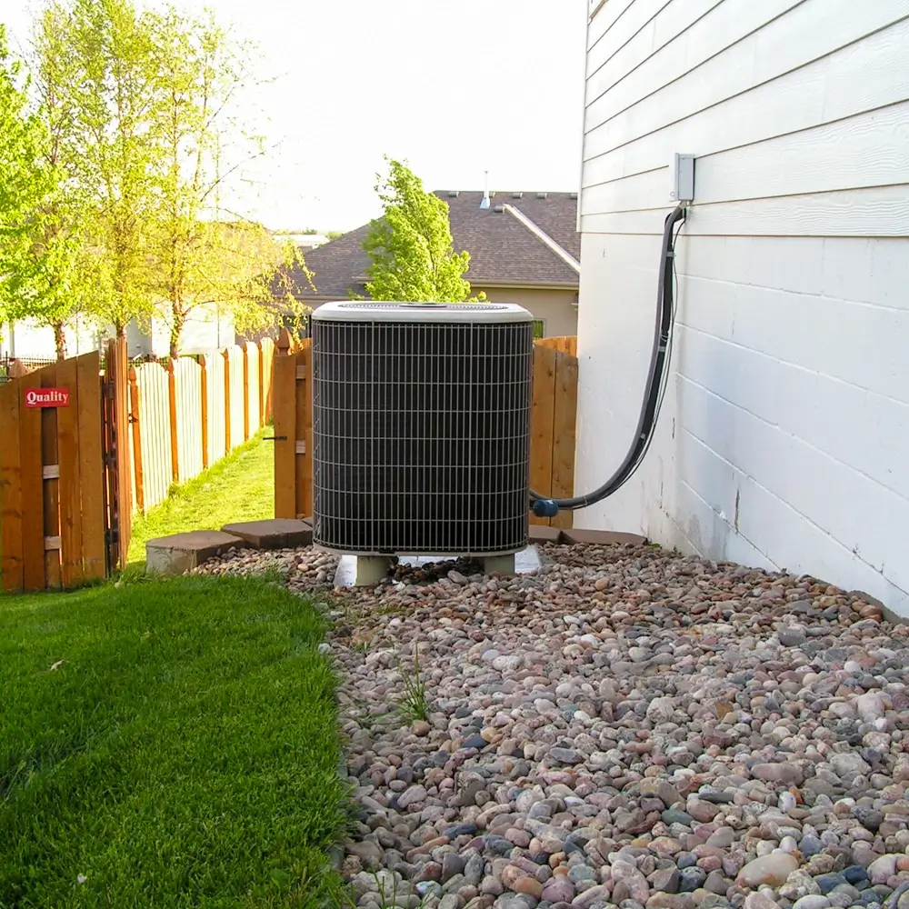 An outdoor air conditioning unit sits on a bed of rocks next to the exterior wall of a white house, with a fenced backyard, green grass, and trees visible in the background.