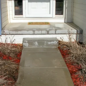A concrete walkway rises steeply to a front porch with no step, creating an awkward slope. Dry plants and red mulch line both sides of the path, leading to a white door with a doormat.