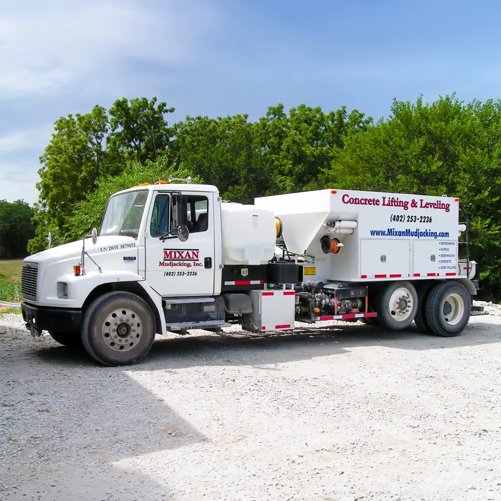 A white Mixan Mudjacking, Inc. truck for concrete lifting and leveling is parked on a gravel lot, with trees and greenery in the background. The truck displays company contact information and equipment on its flatbed.