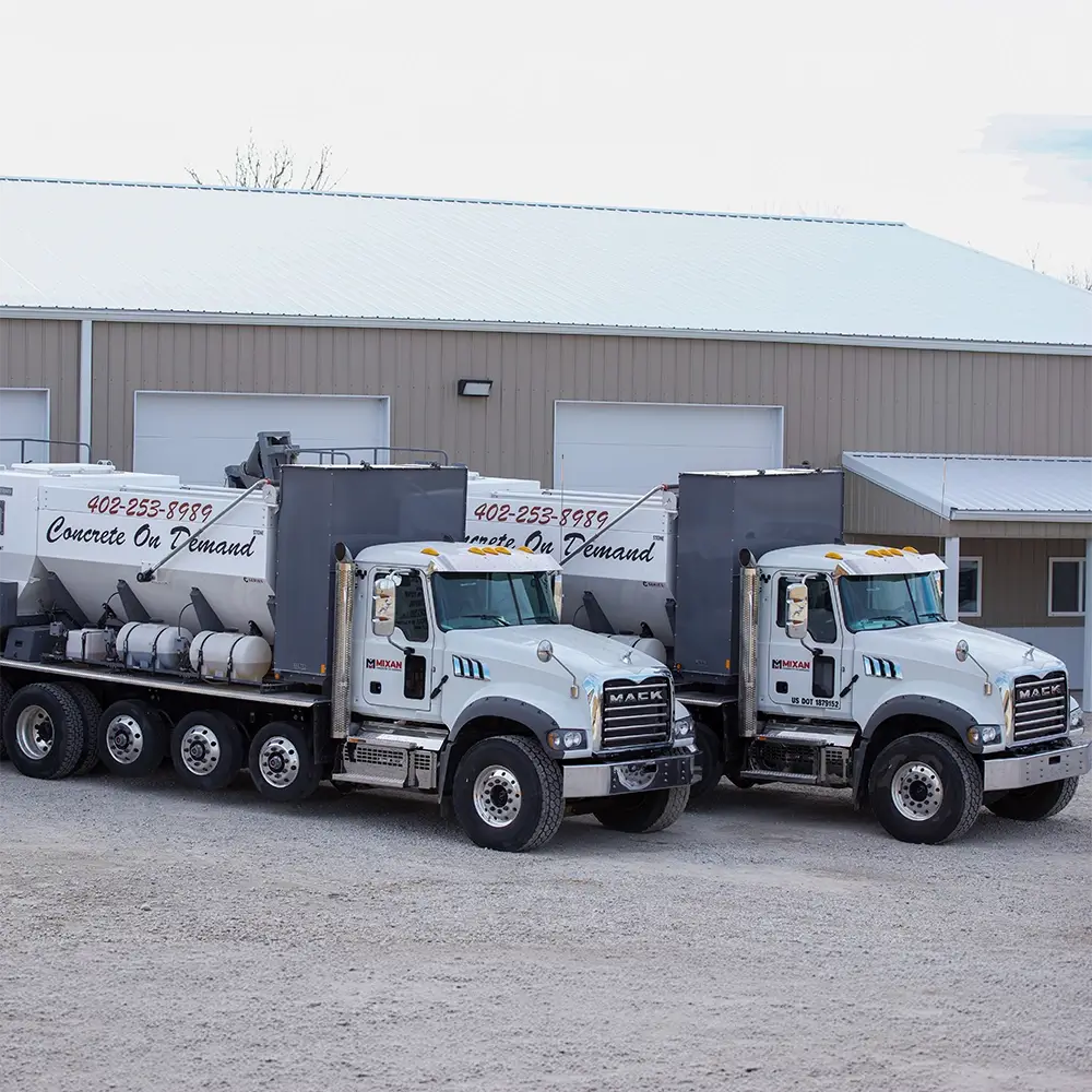 Two white cement mixer trucks labeled Concrete On Demand are parked side by side on a gravel lot in front of a beige industrial building with large garage doors.