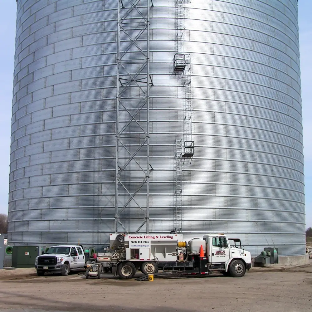 A large cylindrical metal storage tank with a vertical ladder and safety cages stands behind two parked trucks labeled Concrete Lifting & Leveling on a dirt surface.