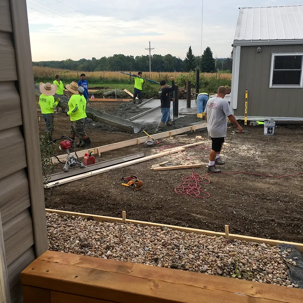 A group of workers in bright shirts are pouring and smoothing concrete for a new sidewalk beside a small building, with tools and construction materials scattered around the worksite. - Mixan Mudjacking A group of workers in bright shirts are pouring and smoothing concrete for a new sidewalk beside a small building, with tools and construction materials scattered around the worksite.