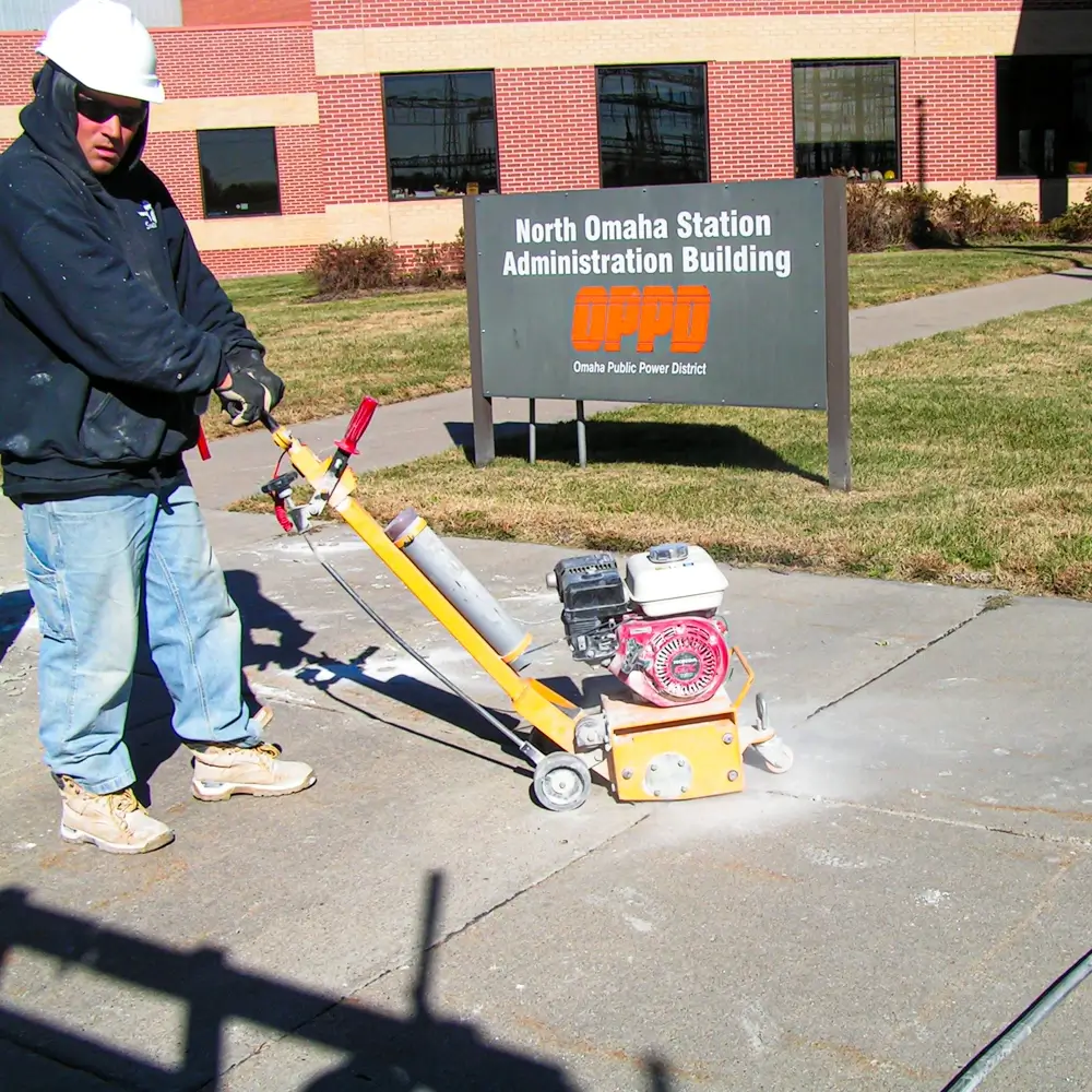 A construction worker wearing a hoodie, hard hat, and sunglasses operates a concrete grinder on a sidewalk near the North Omaha Station Administration Building sign.