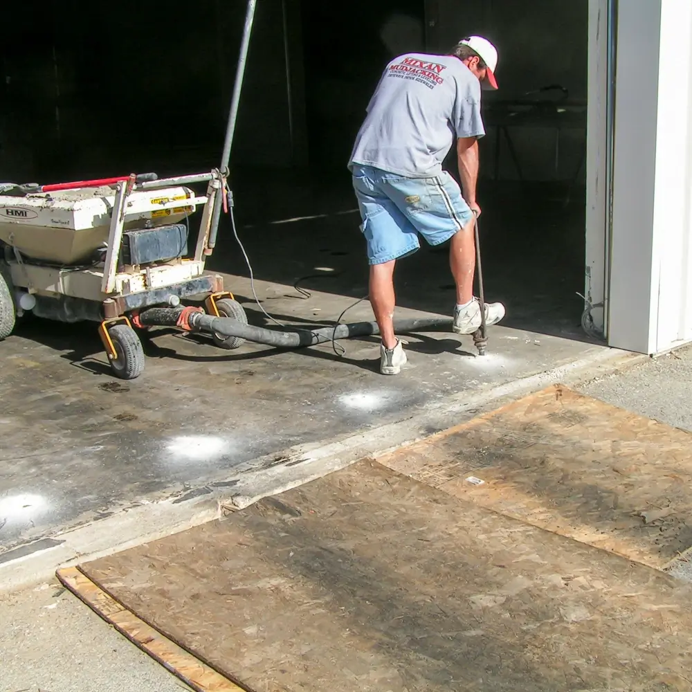 A worker in shorts and a T-shirt uses a long tool to fix or level a concrete garage floor near an open garage door, with machinery and plywood sheets visible nearby.